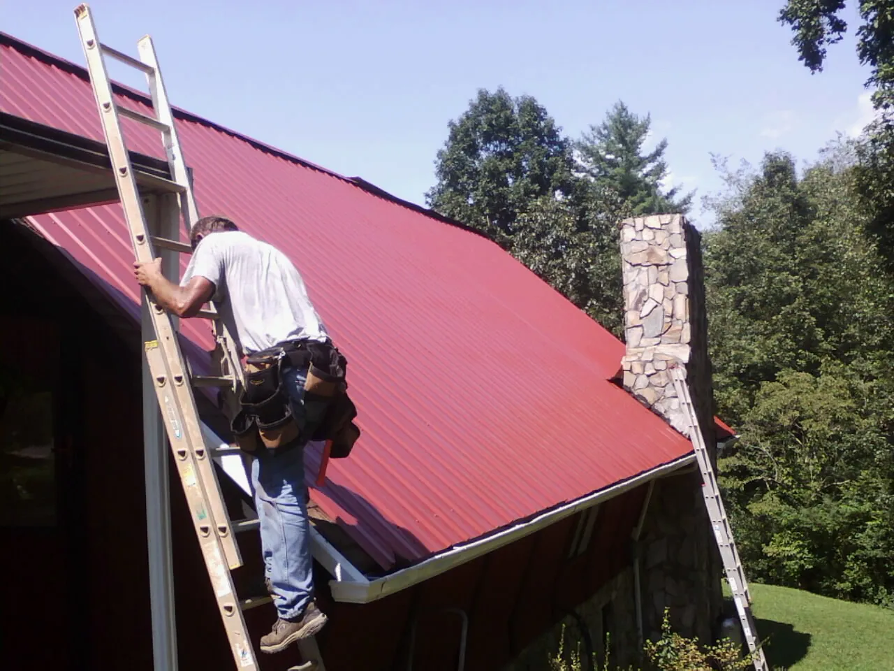 Roofer on a ladder beside a red metal roof and stone chimney — Soffit & Fascia Repair by Roofing Brothers in Barrington